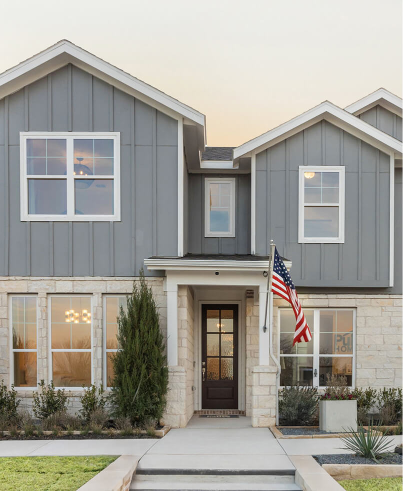 A two-story modern Ramble home with gray siding, stone accents, a front porch, and an American flag displayed by the entrance—ideal for those seeking new homes in DFW or lake homes in North Dallas.