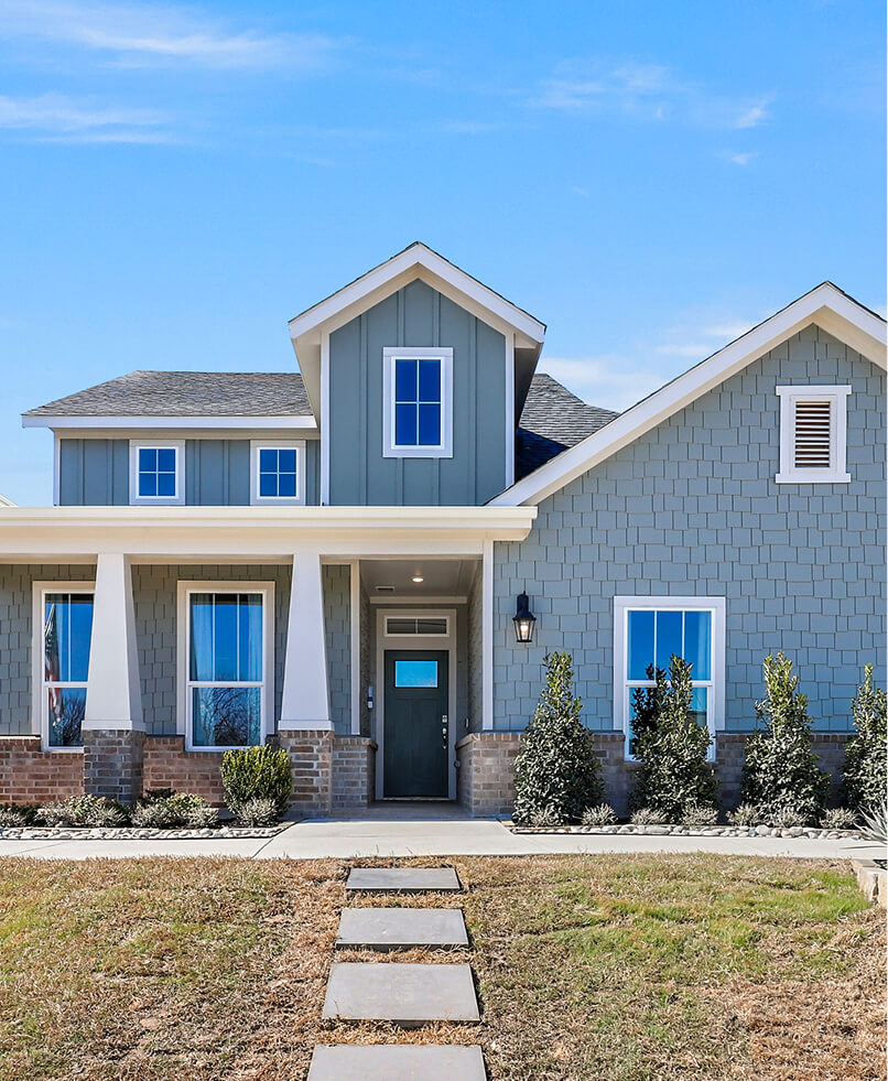 Front view of a modern two-story Ramble home with blue-gray siding, a covered porch, white trim, and a paved walkway leading to the door. Shrubs line the front—perfect for those seeking new homes in Celina TX.