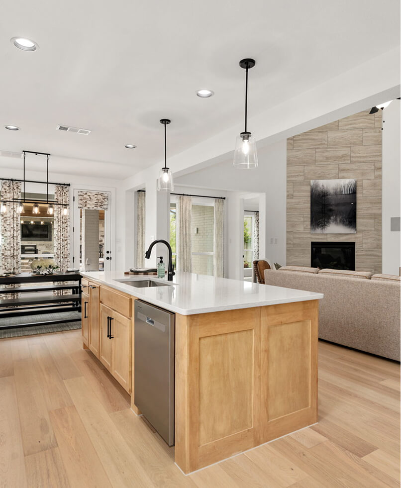 Modern kitchen with a light wood island, white countertops, stainless steel appliances, and pendant lights, opening into a living area with a fireplace and large windows—perfect for those seeking elegant lake homes in North Dallas.