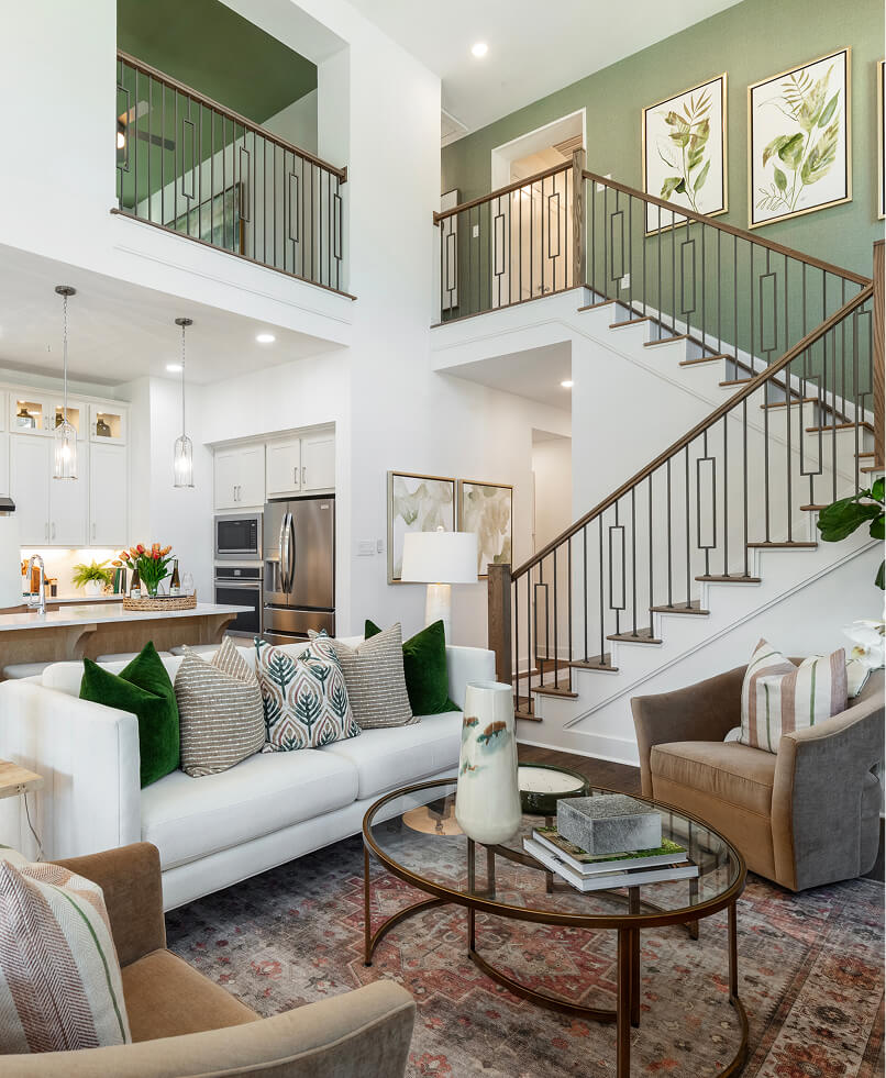 Modern living room with white sofa, green and beige pillows, round glass coffee table, armchair, open kitchen, and staircase with metal railing; botanical artwork on green accent wall—perfect for new homes in North Texas.
