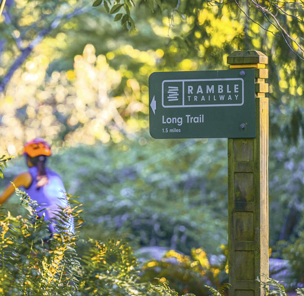 A green signpost reads "Ramble Trailway: Long Trail, 1.5 miles" with a hiker in the background surrounded by greenery—perfect for those seeking lake homes North Dallas or exploring new homes North Texas.