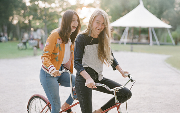 Two young women ride a tandem bicycle together on a gravel path outdoors near beautiful lake homes North Dallas, smiling and enjoying a sunny day. A white tent and trees are visible in the background.
