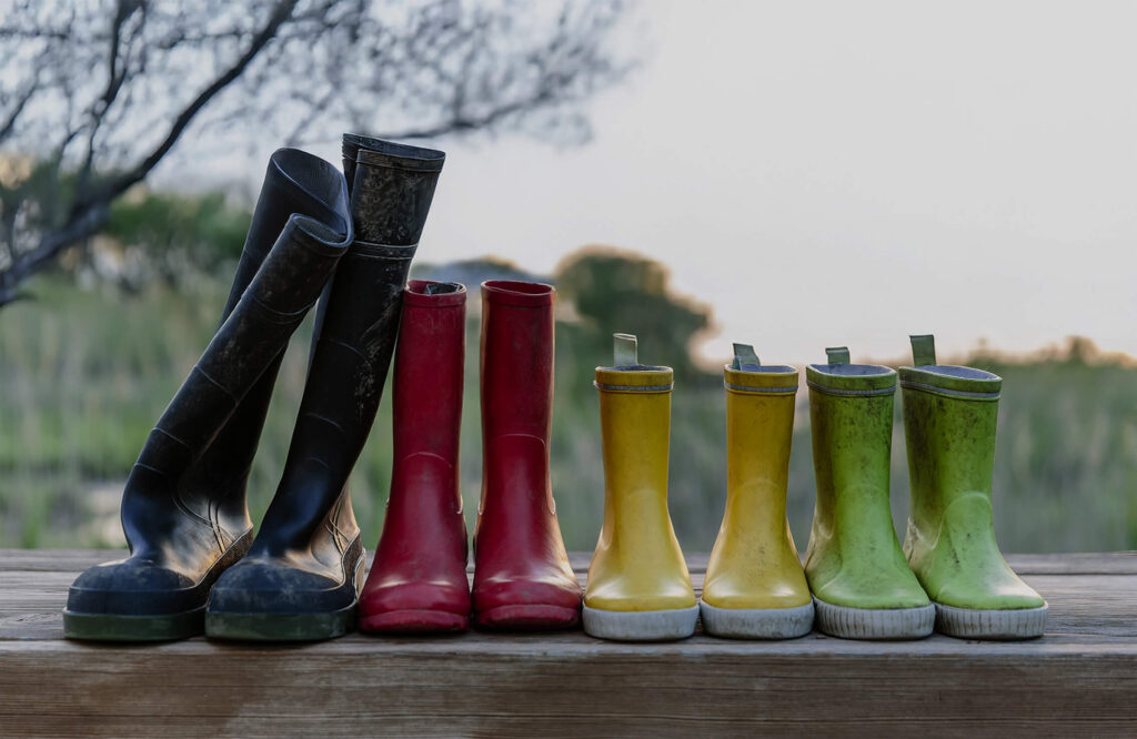 Four pairs of rubber boots—black, red, yellow, and green—are lined up on a wooden surface outdoors with trees in the background, evoking the welcoming spirit of new homes in North Texas.