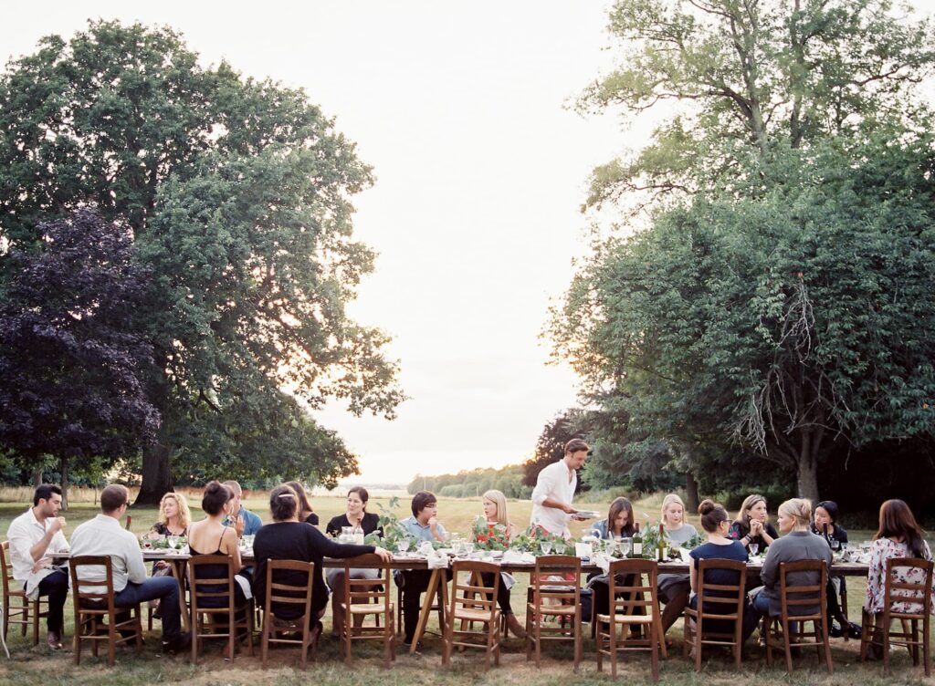 A group of people sit around a long table outdoors, sharing a meal in a grassy area surrounded by trees at sunset—enjoying the peaceful community atmosphere found near Ramble homes and New Homes DFW.