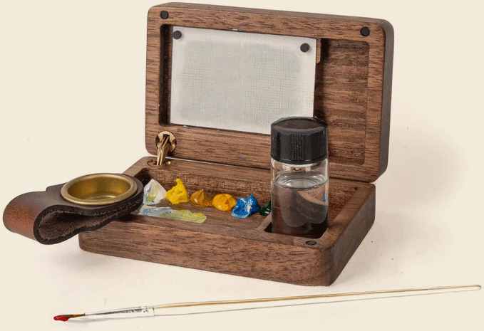 A compact wooden paint box with metal mixing palette, paint blobs, a small jar, a brass cup, and a paintbrush placed in front—perfect inspiration for artists dreaming of designing new homes in North Texas.