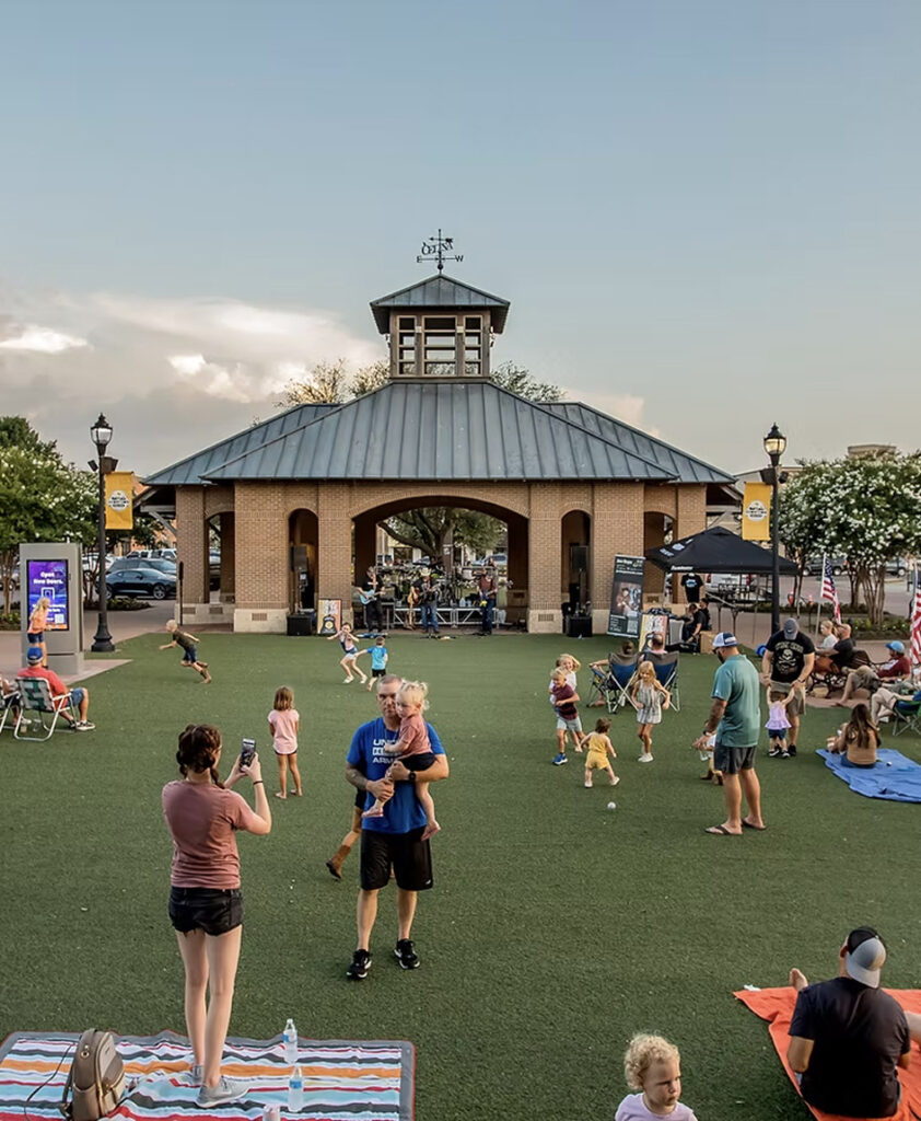 People gather on a grassy lawn in front of a pavilion, with children playing and adults socializing on a clear day—a perfect scene from the vibrant community life found near new homes in North Texas.