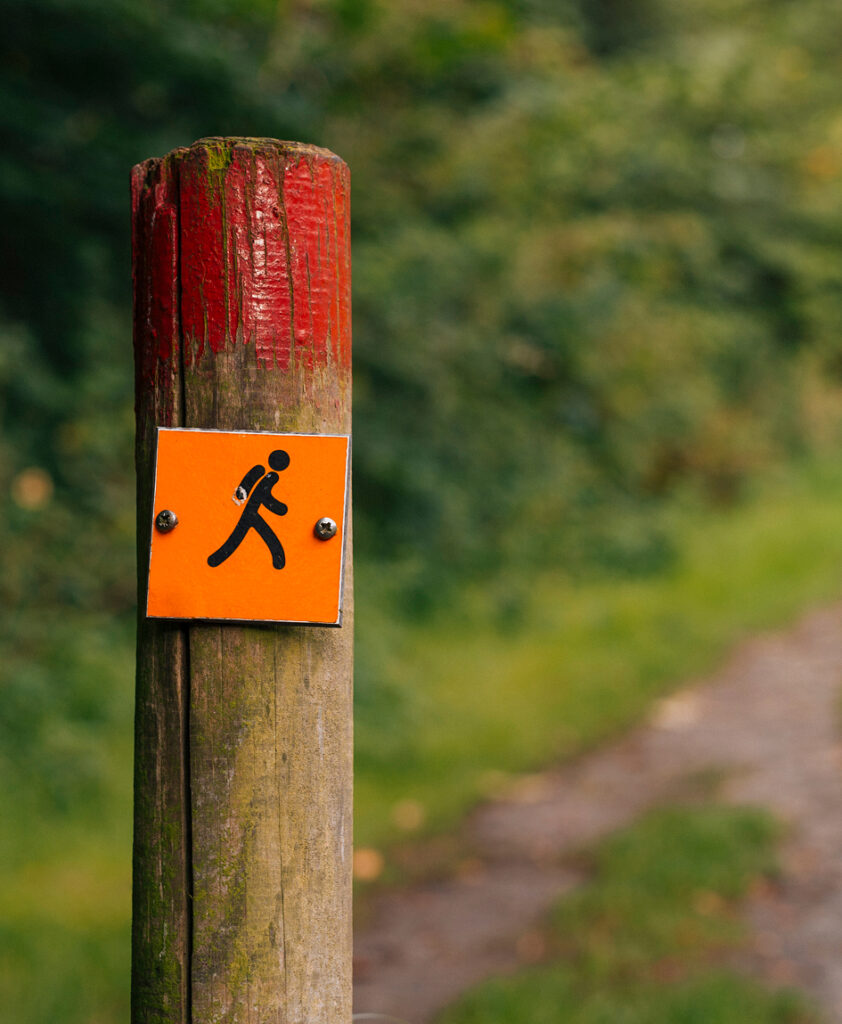 A wooden post with a red top features an orange sign showing a hiker symbol, marking a walking trail next to new homes in Celina, TX, with a blurred path and greenery in the background.