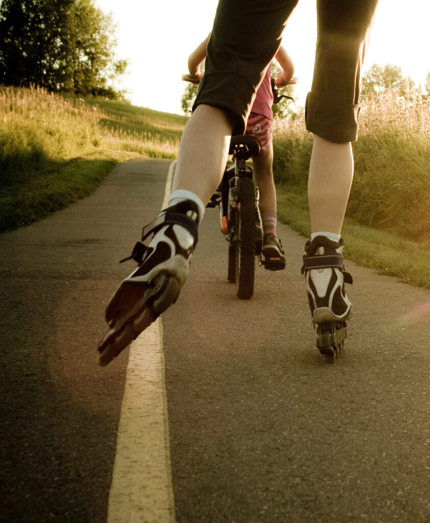 A person on rollerblades and a child on a bicycle ride together on a paved path outdoors in the sunlight, enjoying the scenic surroundings of Ramble homes.