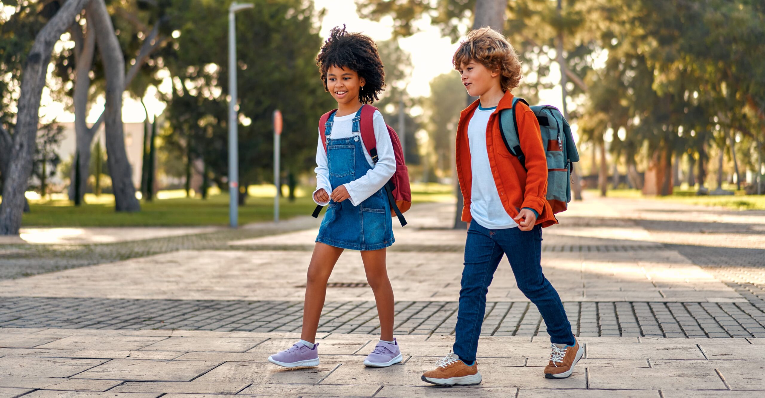 Two young children with backpacks walk side by side on a paved path in a park, surrounded by trees and greenery—capturing the charm of life near new homes in Celina, TX.