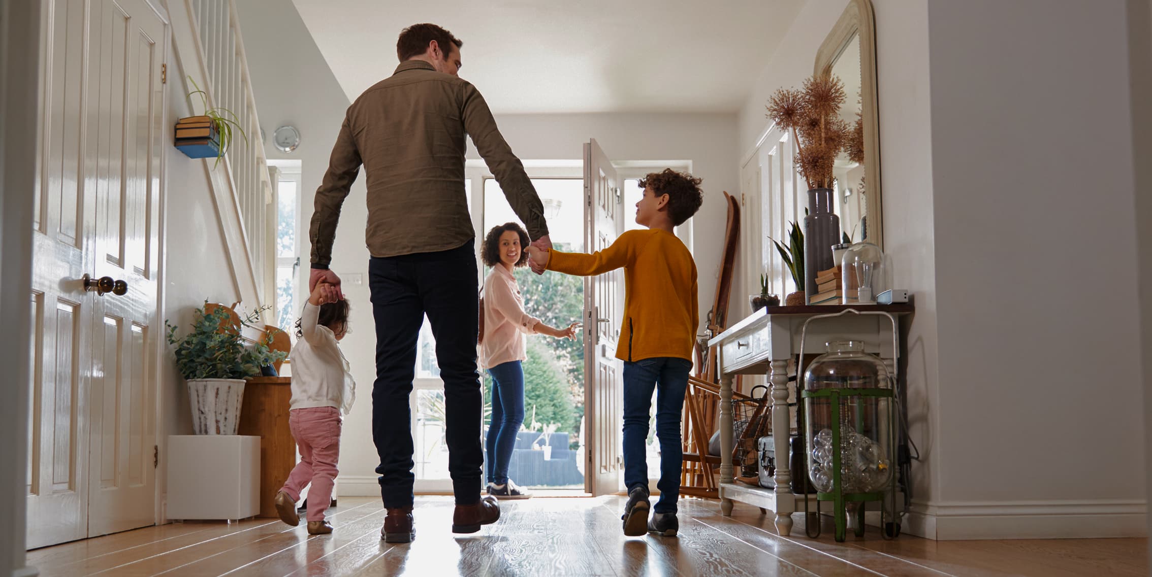 A man stands in a doorway holding a baby while two young children in hats walk onto the porch of one of the beautiful new homes DFW offers, basking in the sunny day.