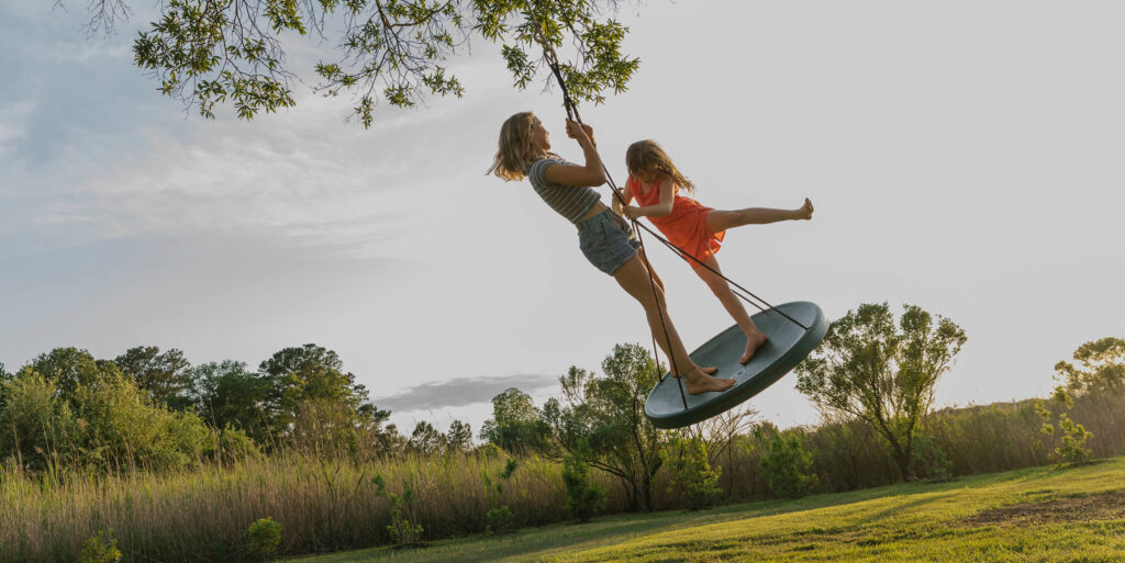 Two girls are playing on a round swing hanging from a tree in the grassy backyard of Ramble homes, with trees and shrubs providing a scenic backdrop—perfect for families seeking new homes in North Texas.