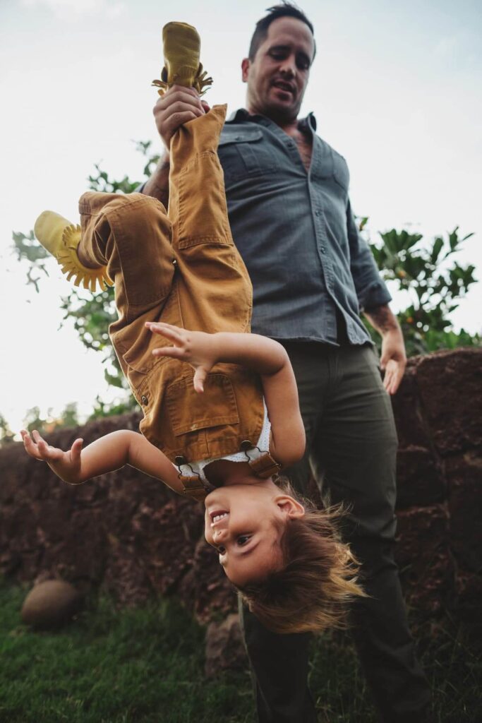 An adult holds a smiling child upside down by the ankle outdoors, with trees and a stone wall in the background—a joyful moment near beautiful lake homes North Dallas.