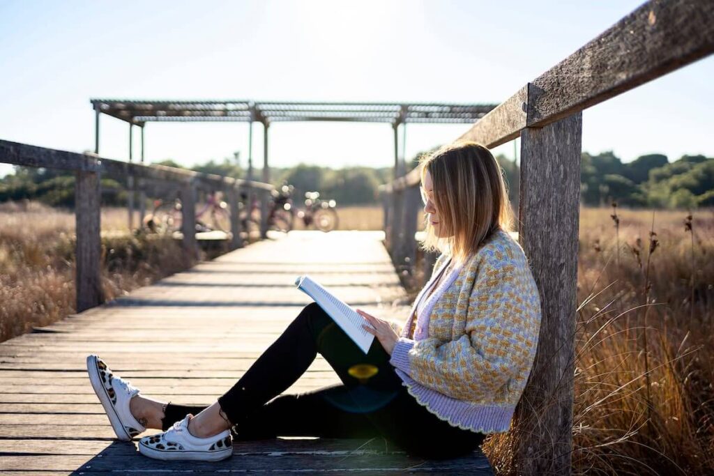 A woman sits on a wooden walkway in sunlight, reading a book with tall grass and distant trees in the background—capturing the serenity often found near Lake homes North Dallas.