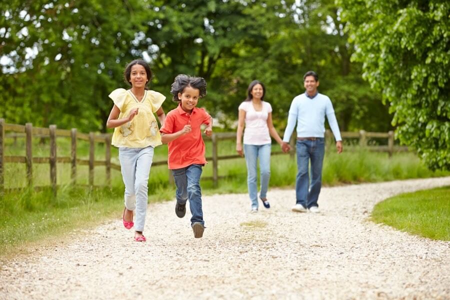 Two children run ahead on a gravel path while two adults walk behind them in a park setting with trees and a wooden fence, capturing the family-friendly atmosphere found near new homes in Celina, TX.