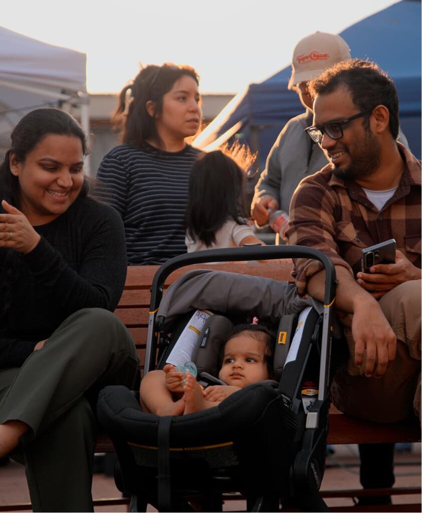 A group of adults sit on benches outdoors, smiling and talking, while a baby rests in a car seat in the foreground.