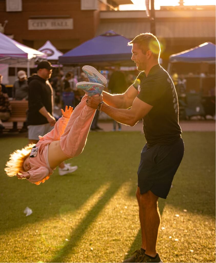 An adult swings a child by the arms in an outdoor event area, with tents, people, and sunlight in the background.