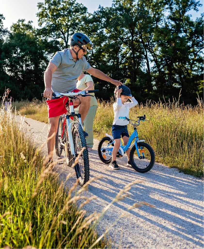 An adult helps a child adjust their helmet while both stand with bikes on a gravel path surrounded by tall grass and trees.