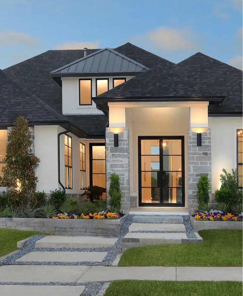 Modern two-story house with large windows, light-colored stone and stucco exterior, black roof, landscaped front yard, and illuminated lights by the entrance.