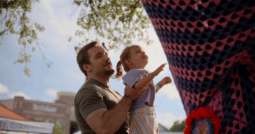 A man holds a young child who is reaching towards a large, colorful crocheted installation at an outdoor event.
