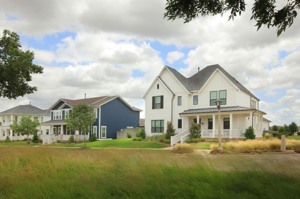 Row of modern suburban houses with well-kept lawns, trees, and cloudy sky in the background. The homes have white and blue exteriors with large windows and front porches.