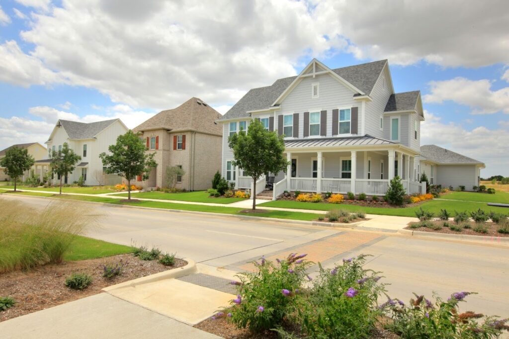 Suburban street with modern two-story houses, well-kept lawns, small trees, and flowerbeds under a partly cloudy sky.