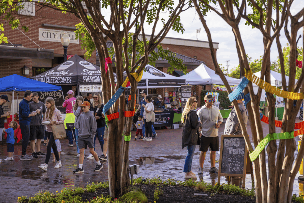 People visit vendor booths at an outdoor market or festival, with colorful decorations on trees and a sign for free summer events visible in the foreground.